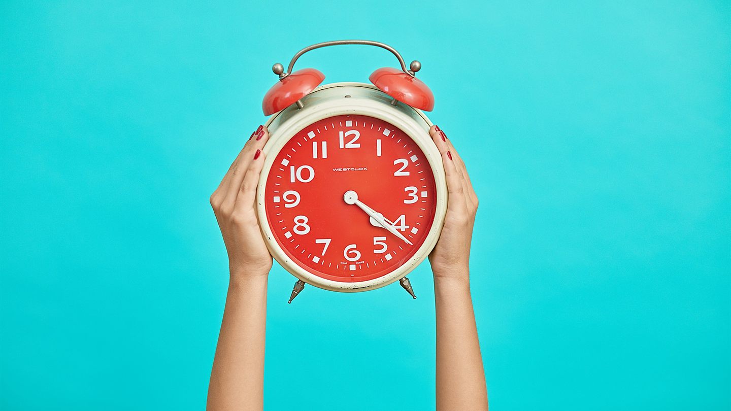Hands holding a red alarm clock overhead against a turquoise background