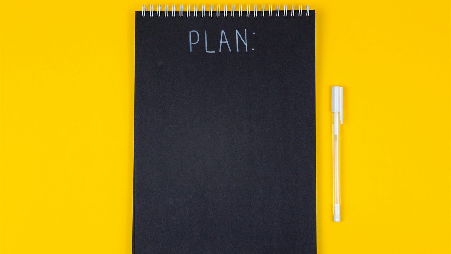 Чёрный блокнот с надписью PLAN и белая ручка на жёлтом фоне